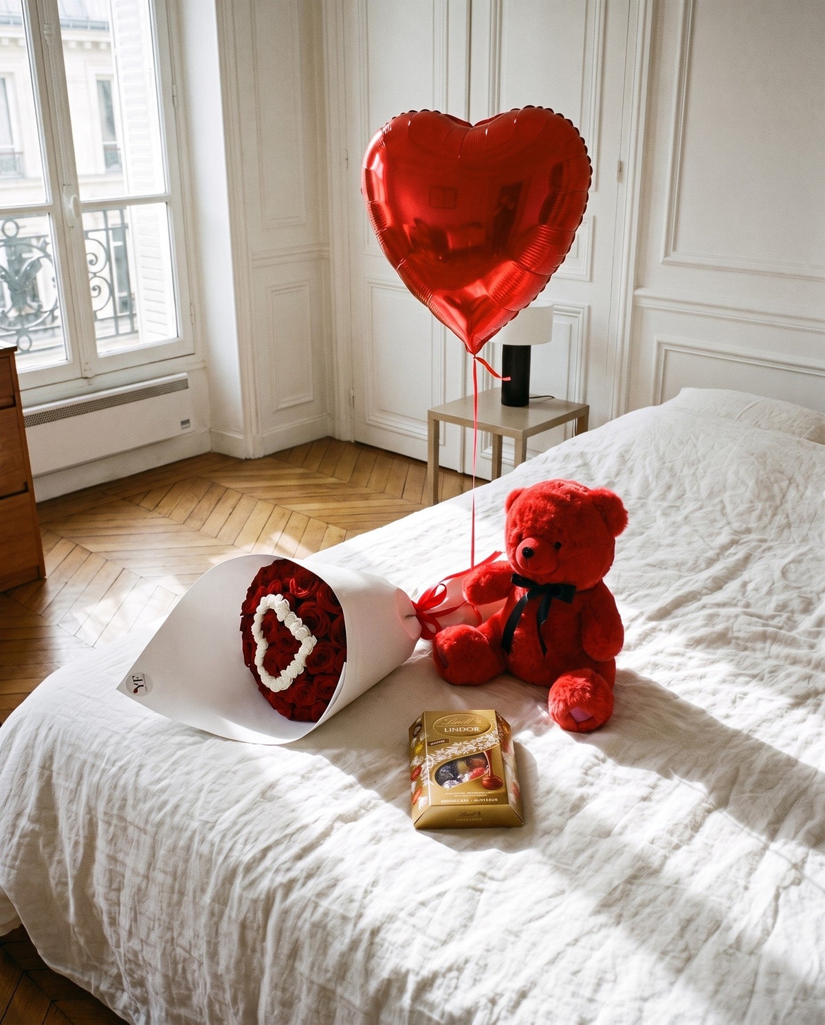 Un bouquet de roses au cœur blanc, un nounours rouge, des chocolats et un ballon cœur réunis sur un lit